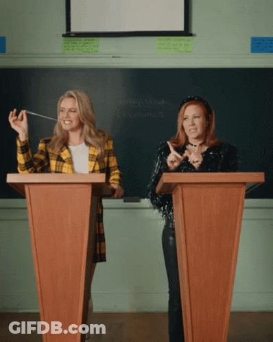Two Women At Podiums With Economics Chalkboard GIF
