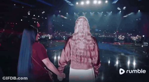 Two Women Holding Hands In Front Of A Stage With A Rumble Logo In The Corner GIF