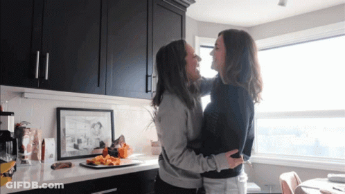Two Women Hugging In A Kitchen With Black Cabinets And A Picture On The Wall GIF