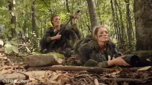Two Women In Camouflage Are Laying In The Woods Holding Guns . GIF