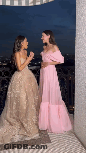 Two Women In Long Dresses Are Standing Next To Each Other On A Balcony At Night GIF