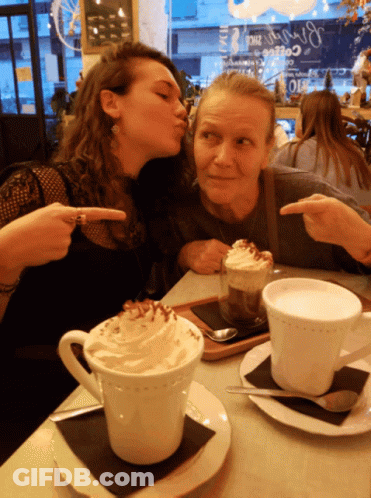 Two Women Kissing On Cheek At Table With Coffee GIF