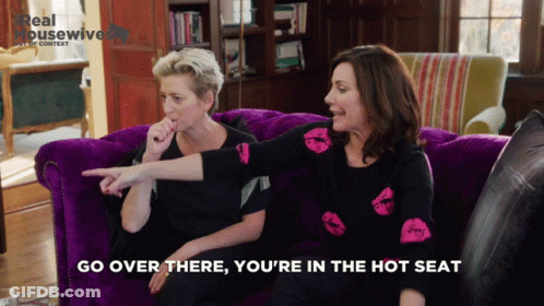 Two Women On Purple Couch Go Over There You're In The Hot Seat GIF