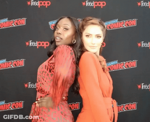 Two Women Pose For A Photo In Front Of A Comic Con Backdrop GIF