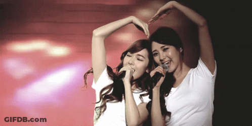 Two Women Singing Into Microphones And Making A Heart Shape GIF