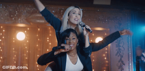 Two Women Singing Into Microphones With One Sitting On The Other 'S Shoulders GIF
