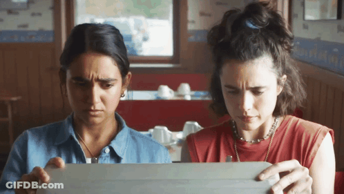 Two Women Sit At A Table Looking At A Box GIF