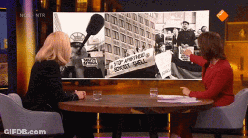 Two Women Sitting At A Table In Front Of A Screen That Says Nos On It GIF