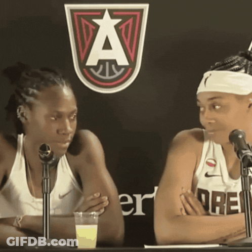 Two Women Sitting At Table With Microphones And Sign A GIF