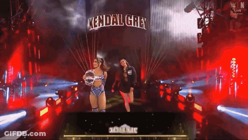 Two Women Standing In Front Of A Sign That Says Kendal Grey On It GIF