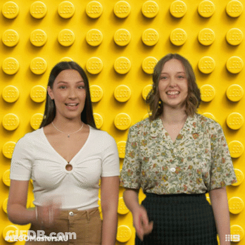 Two Women Standing In Front Of Yellow Lego Wall GIF