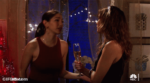 Two Women Standing Next To Each Other Holding A Glass Of Champagne With The Nbc Logo On The Bottom GIF