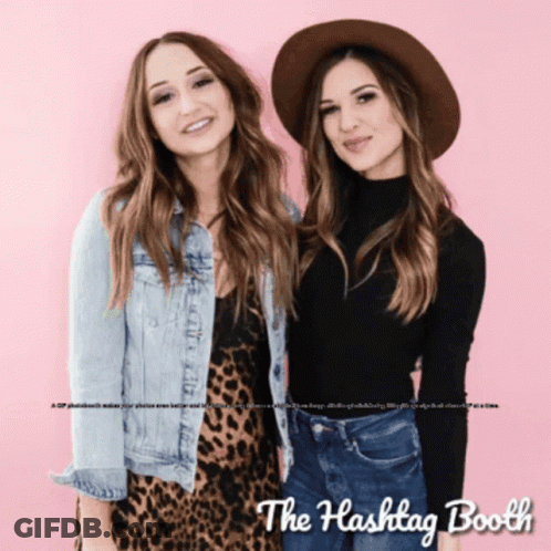 Two Women Standing Next To Each Other In Front Of A Pink Background With The Hashtag Booth Written On It GIF