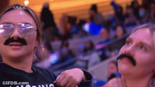Two Women Wearing Fake Mustaches And Sunglasses Are Standing In A Crowd .. GIF