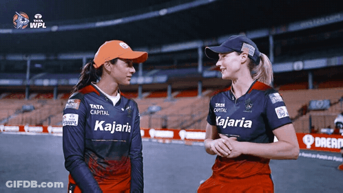 Two Women Wearing Kajaria Shirts Are Standing Next To Each Other On A Field GIF