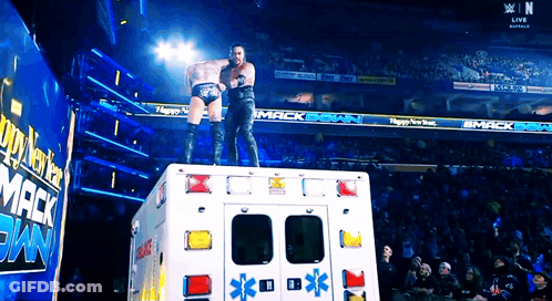 Two Wrestlers Are Standing On Top Of An Ambulance In Front Of A Sign That Says Mack Bank GIF