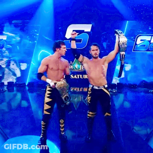 Two Wrestlers Holding Up Their Championship Belts In Front Of A Saturn Sign GIF