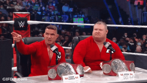Two Wrestlers Sitting At A Table With A Sign That Says Chad Gable On It GIF