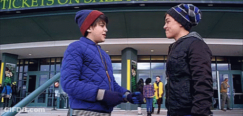 Two Young Boys Are Shaking Hands In Front Of A Building That Says Tickets On It GIF