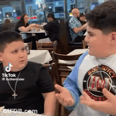 Two Young Boys Are Sitting At A Table In A Restaurant Talking . GIF