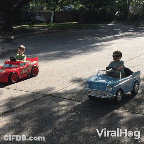 Two Young Boys Driving Toy Cars On Street GIF