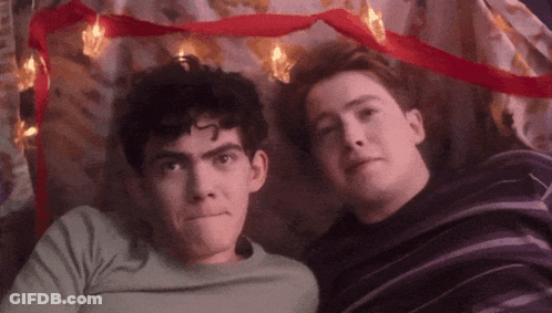 Two Young Men Are Laying Next To Each Other Under A Blanket With Christmas Lights . GIF