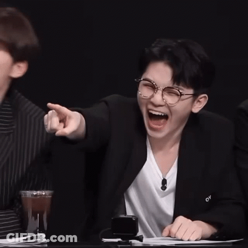 Two Young Men Are Sitting At A Table Laughing And Pointing At Something . GIF
