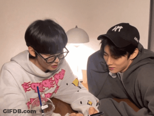 Two Young Men Are Sitting At A Table One Wearing A Hat That Says Ny GIF