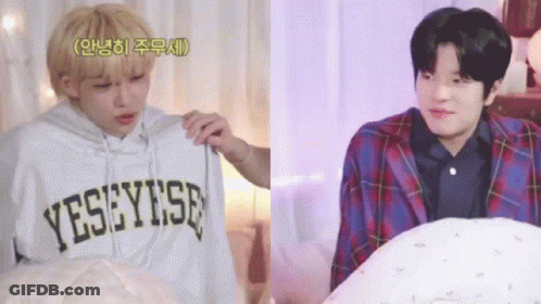 Two Young Men Are Sitting Next To Each Other In A Bedroom . One Of The Men Is Holding A Hoodie . GIF