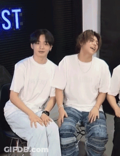 Two Young Men Are Sitting Next To Each Other In Front Of A Neon Sign That Says St GIF