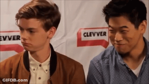 Two Young Men Are Sitting Next To Each Other In Front Of A Sign That Says ' Clever ' On It . GIF