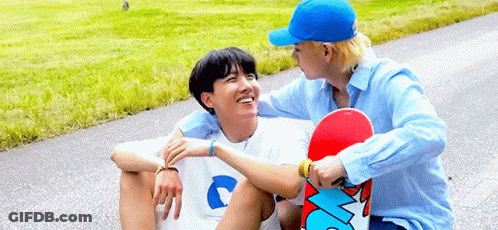 Two Young Men Are Sitting On The Side Of A Road Holding A Skateboard . GIF