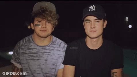 Two Young Men Are Standing Next To Each Other In A Dark Room . One Of The Men Is Wearing A Ny Hat . GIF