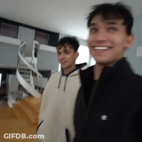 Two Young Men Are Standing Next To Each Other In A Room With A Spiral Staircase In The Background . GIF