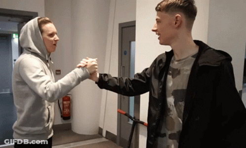Two Young Men Shake Hands In A Hallway With A Fire Extinguisher On The Wall GIF