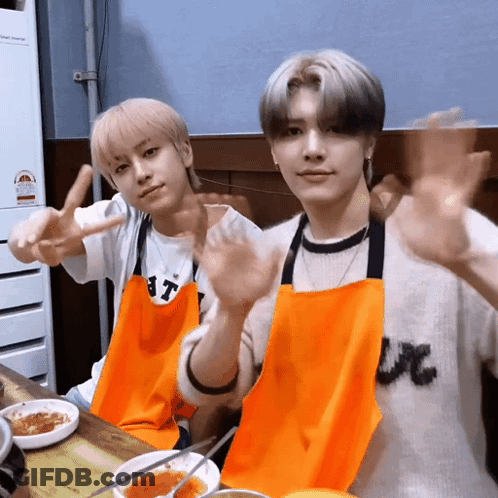 Two Young Men Wearing Aprons Are Sitting At A Table GIF