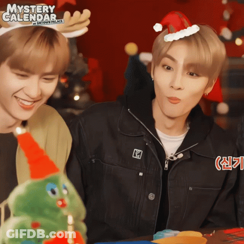 Two Young Men Wearing Santa Hats And Antlers Are Standing Next To A Christmas Tree And A Sign That Says Mystery Calendar GIF