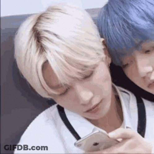Two Young Men With Blonde Hair And Blue Hair Are Sitting Next To Each Other And Looking At A Cell Phone . GIF