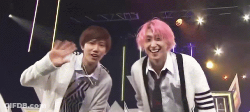 Two Young Men With Pink Hair Are Standing Next To Each Other On A Stage And Waving At The Camera . GIF