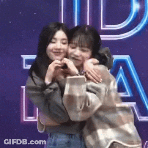 Two Young Women Hugging In Front Of Purple Sign GIF