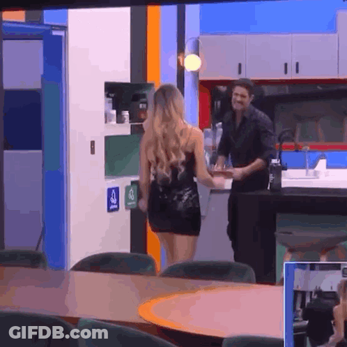 Woman In Black Dress Walking Toward Man In Black Shirt In Kitchen GIF