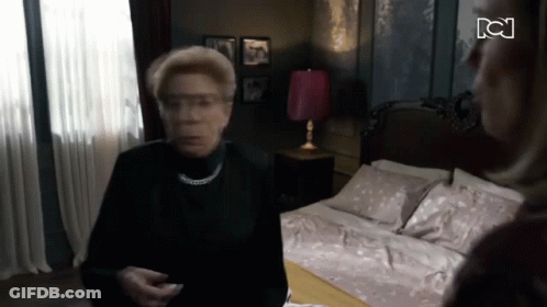 Woman In Black Shirt Standing In Front Of Bed With Pink Lamp GIF