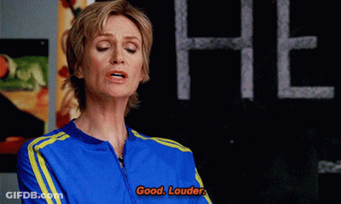 Woman In Blue And Yellow Jacket Says Good Louder In Front Of Chalkboard GIF