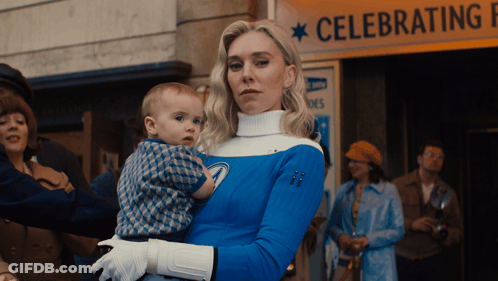 Woman In Blue Costume Holding Baby In Front Of Celebrating Sign GIF