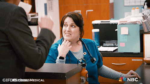 Woman In Blue Scrub Talking To Man In Black Suit With NBC Logo GIF