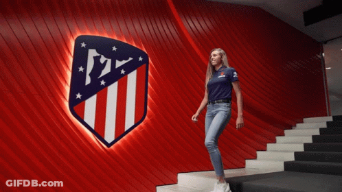 Woman In Blue Shirt Walking Down Stairs In Front Of Atlético Madrid Sign GIF