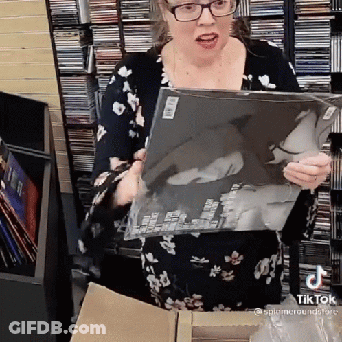 Woman In Floral Dress Holding Record In Front Of CD Shelf GIF