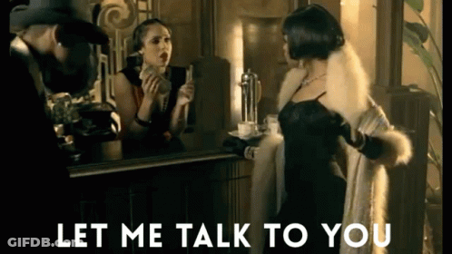 Woman In Fur Coat Talking To Man In Hat In Front Of Mirror GIF