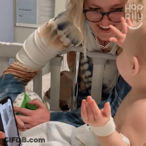 Woman In Glasses And Tie-Dye Shirt Talks To Baby In Hospital Crib GIF