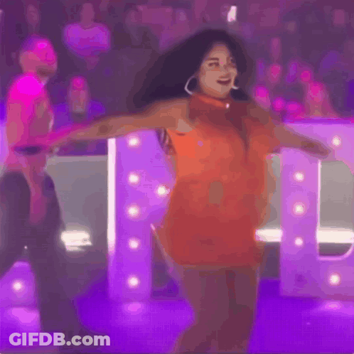 Woman In Orange Dress Dancing In Front Of Purple Letter GIF